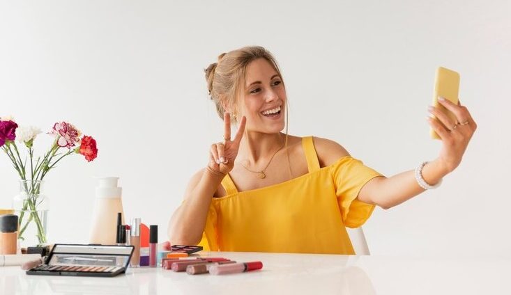 Woman in a yellow top taking a selfie while sitting at a table with various makeup products.